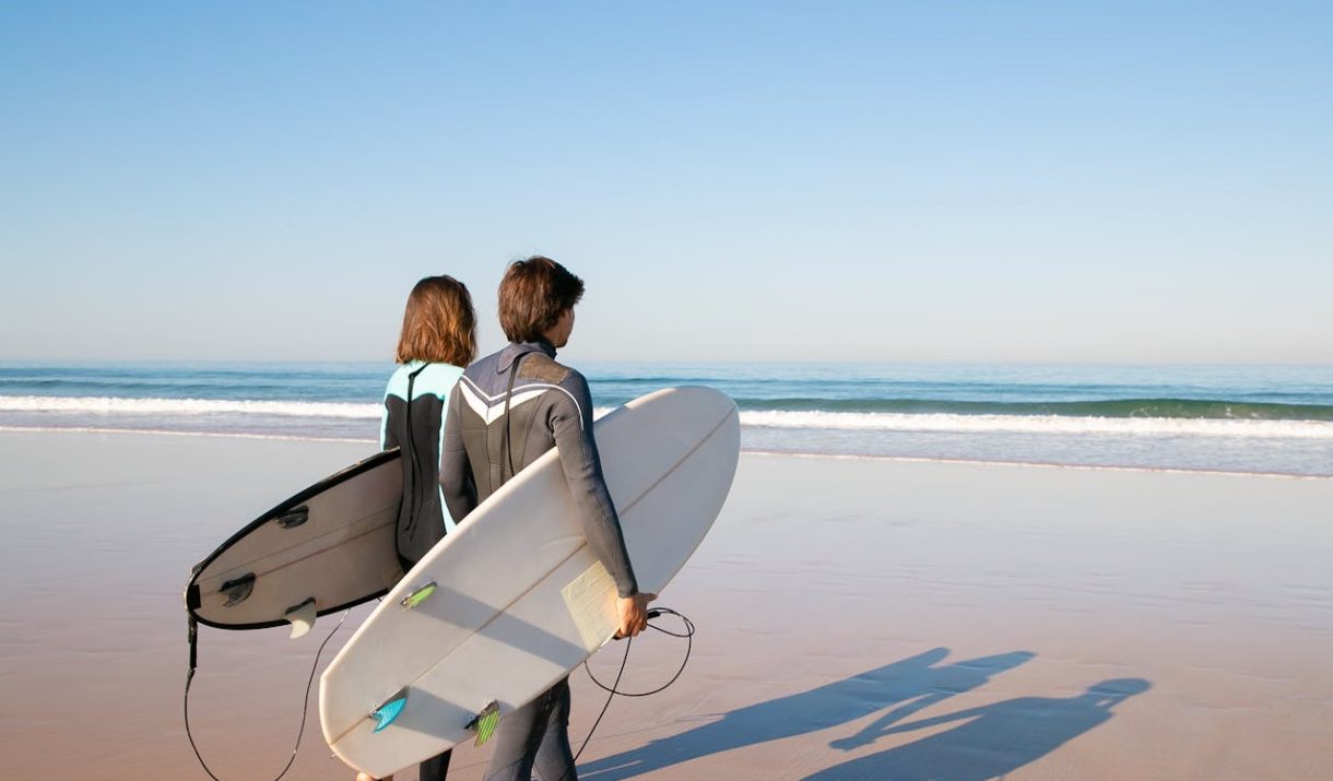 A young couple in wetsuits carrying surfboards along a sunny beach in Portugal.