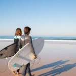 A young couple in wetsuits carrying surfboards along a sunny beach in Portugal.